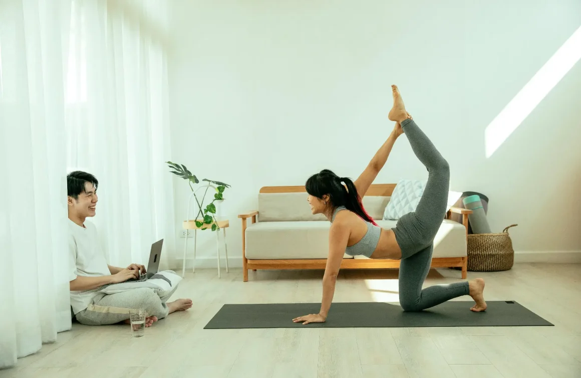 A woman in gray athletic wear performs a high-leg yoga pose on a mat in a bright living room while a man sits cross-legged nearby with a laptop, smiling.