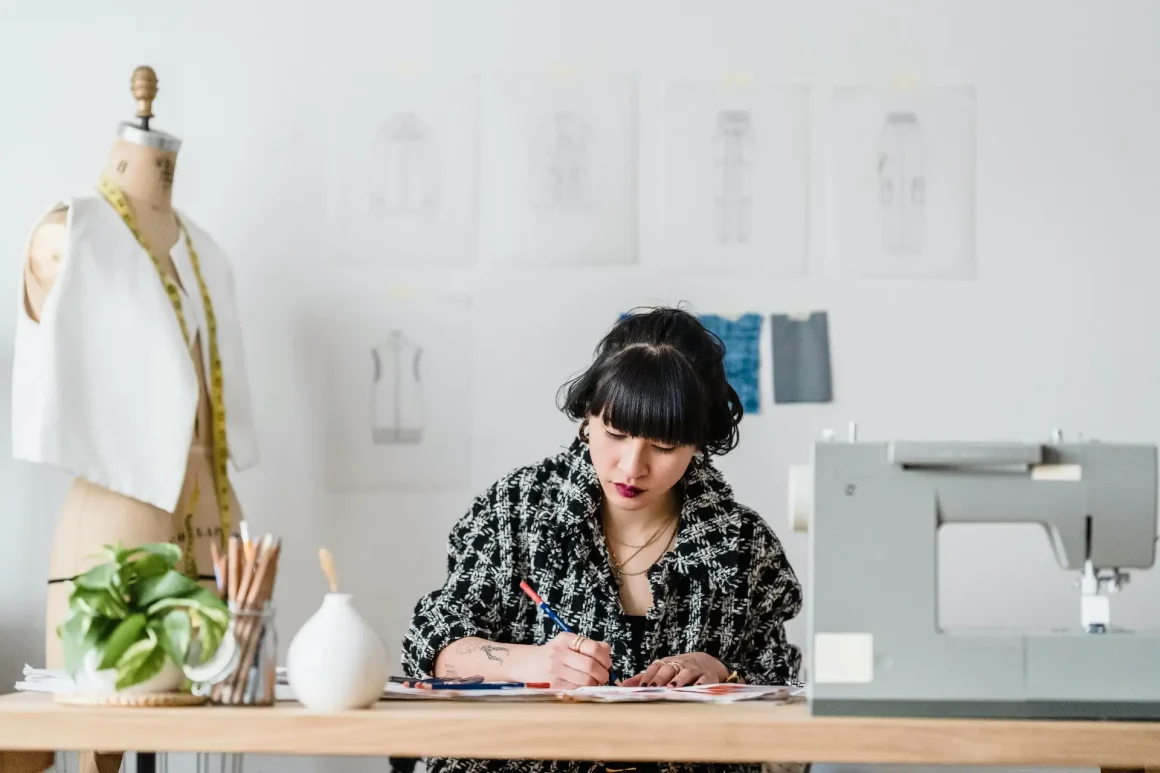 A woman with black hair and bangs sits at a wooden table, drawing with colored pencils. She wears a black and white patterned shirt. A sewing machine, mannequin, plants, and design sketches on the wall populate the light-filled studio.