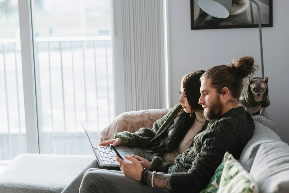 A man and a woman sit together on a gray sofa by a large window, looking at a laptop. The man has a beard and hair in a bun, wearing a dark sweater. The woman wears a green cardigan. A small dog sits behind them, watching, with a framed picture and a modern floor lamp in the background.