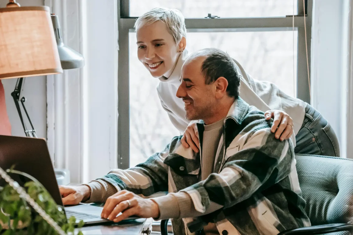 A smiling woman with short blonde hair leans over a man sitting at a desk, her hands resting on his shoulders as they both look at a laptop screen by a window with a warm desk lamp nearby.