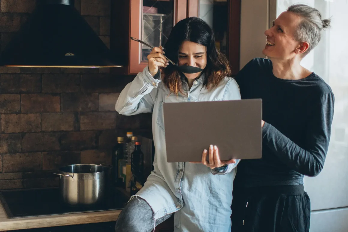 A couple in a kitchen: one woman holding a laptop and playfully placing a spoon under her nose while another woman in a black top smiles beside her, with a pot on the stove and bottles in the background.
