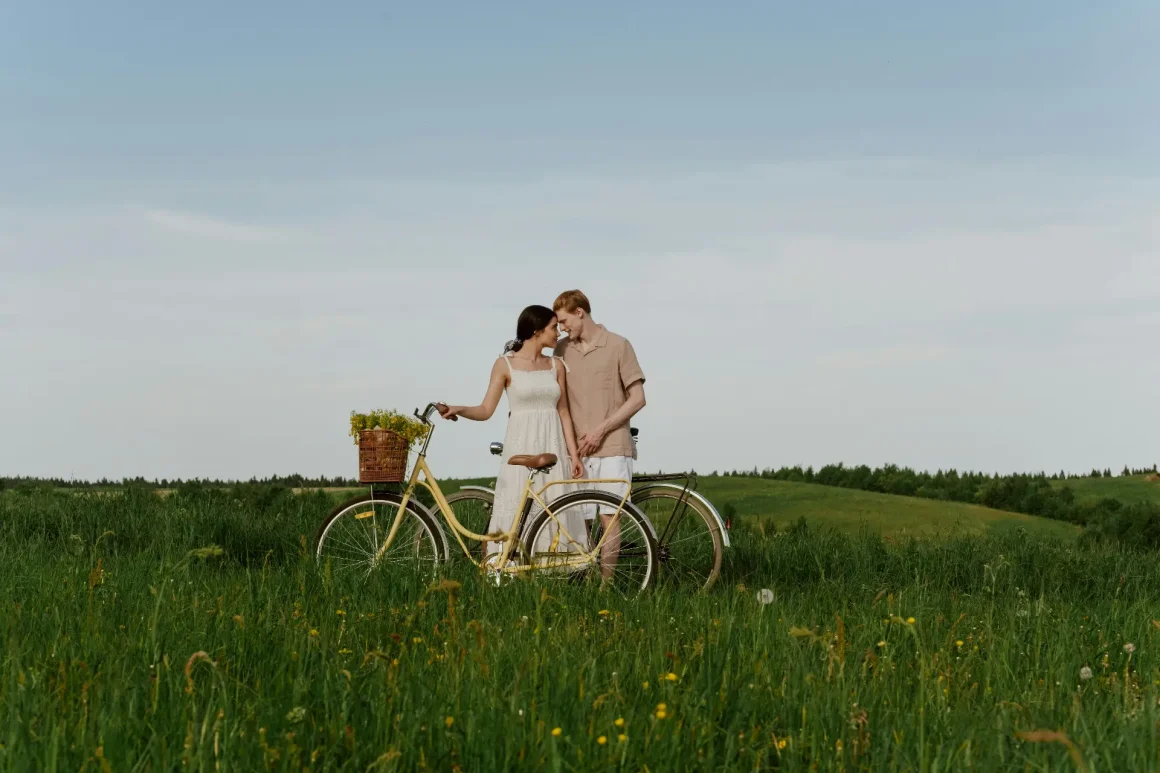 A couple stands in a green meadow beside a vintage yellow bicycle with a wicker basket of flowers. The woman wearing a white dress and the man in a light brown shirt and shorts lean close together, holding hands, under a pale blue sky.
