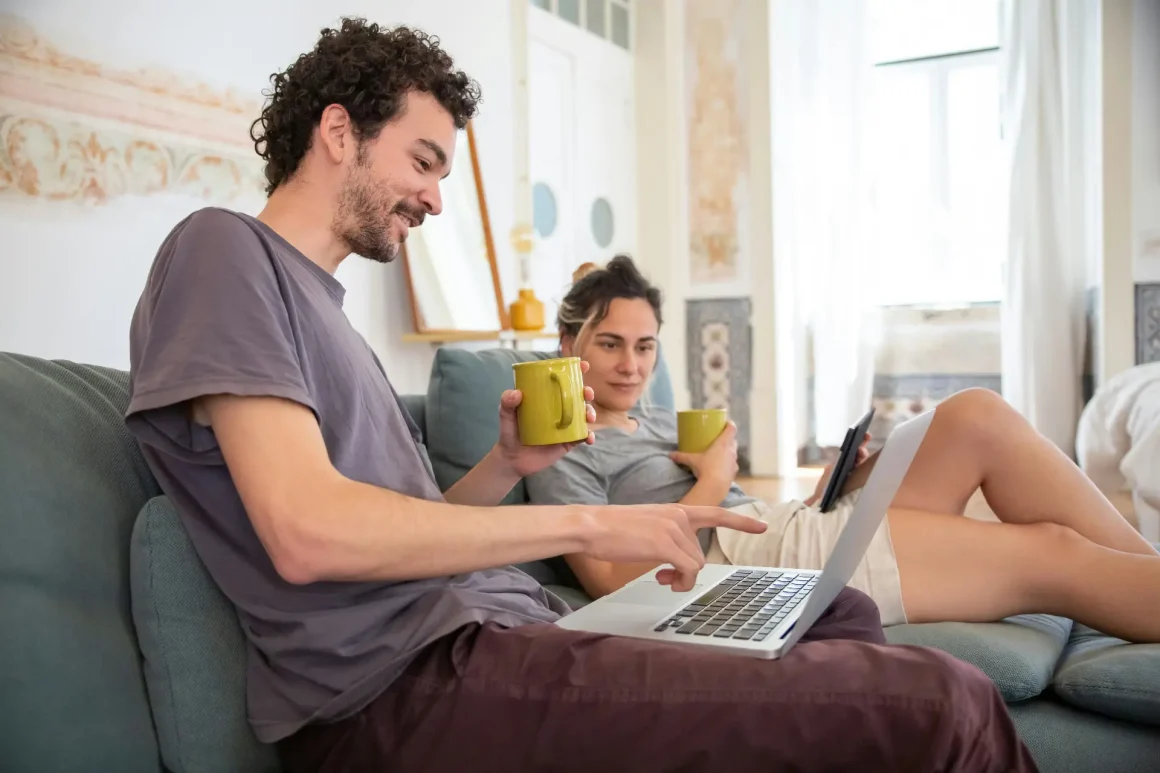A man with curly hair in a gray t-shirt uses a laptop on a teal sofa, while a woman reclining on the same couch holds a yellow mug and a tablet, smiling softly in a bright living room.