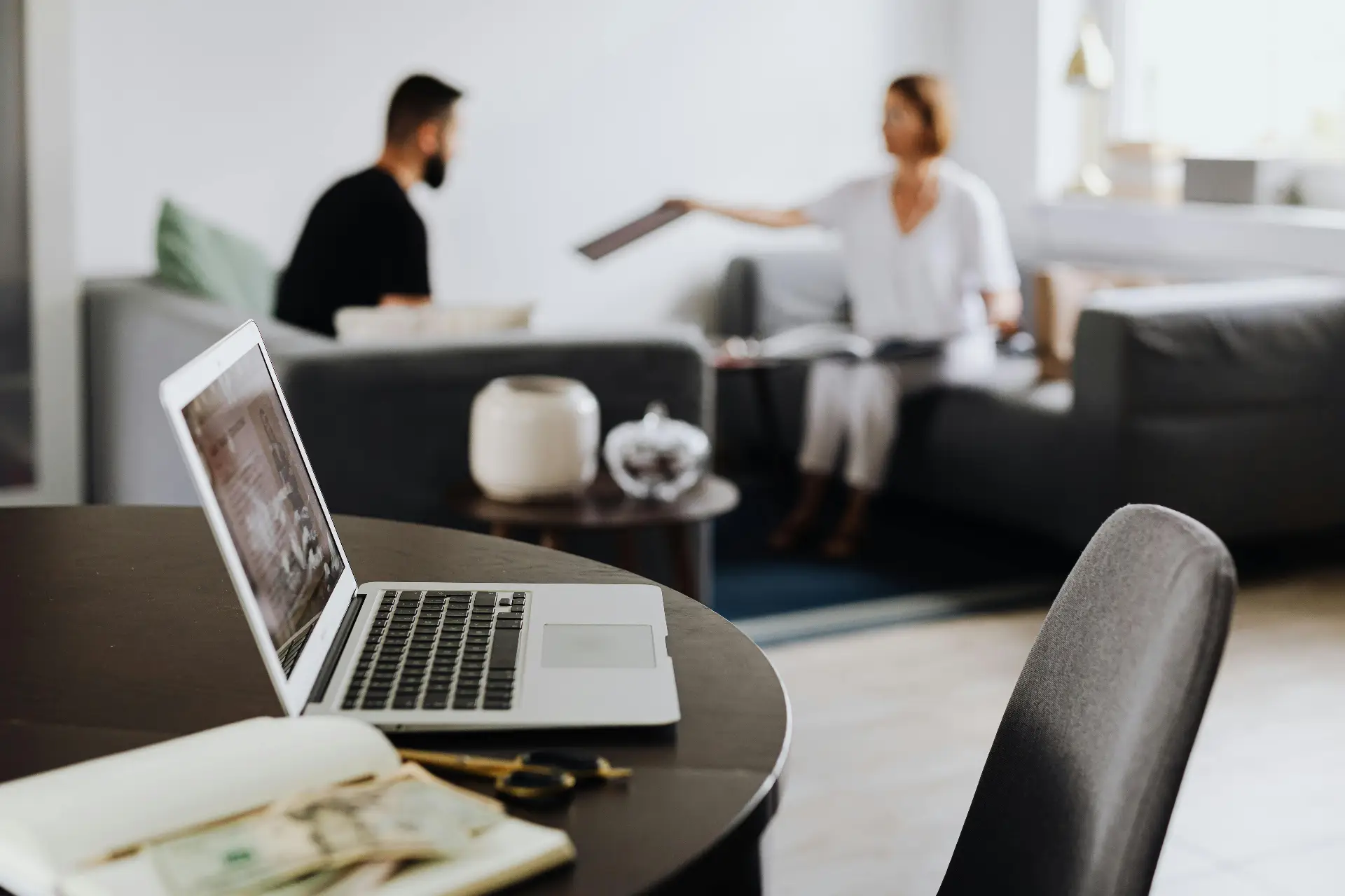Cozy Home Office Meeting Scene A laptop on a round wooden table in the foreground, with two people blurred in the background sitting on a couch and passing a tablet or document between them, in a bright living room setting.