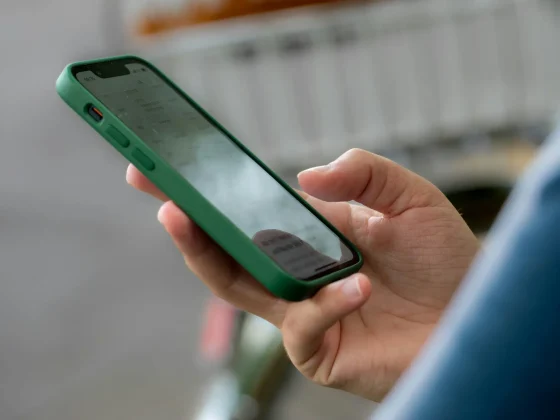 Close-up of a hand holding a smartphone with a green protective case, screen visible, outdoors with a blurred background.