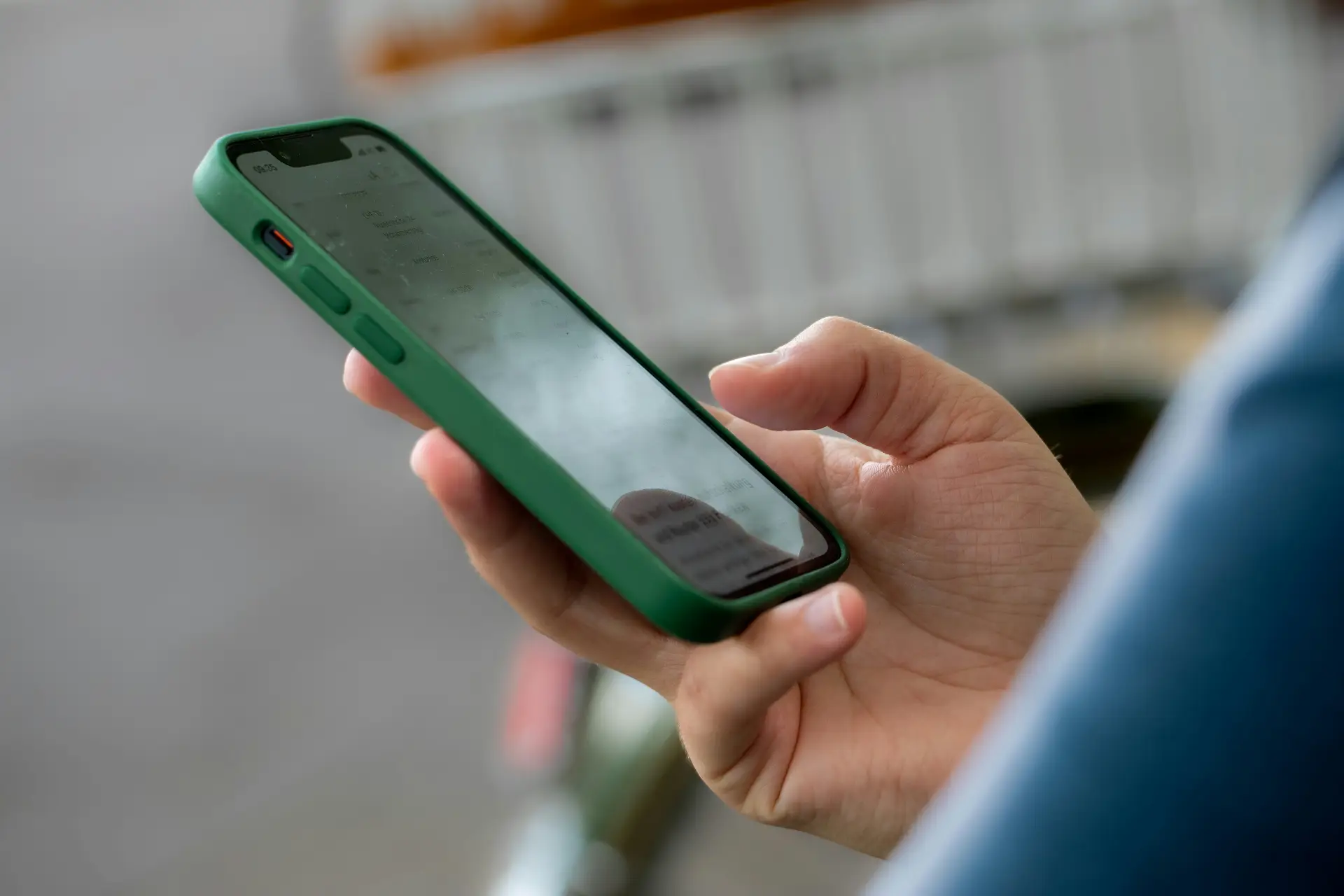 Close-up of a hand holding a smartphone with a green protective case, screen visible, outdoors with a blurred background.