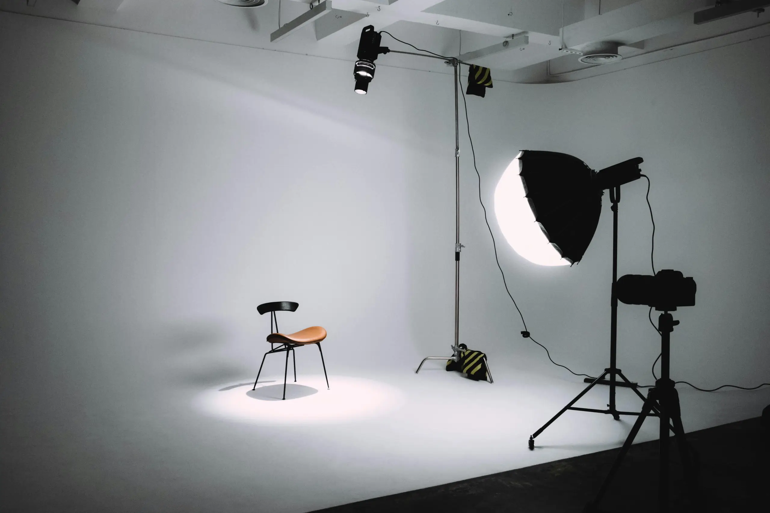 A small brown chair sits on a white photography studio backdrop, illuminated by a bright circular light on the right and a large softbox on a stand. A few cables and stands are visible, with overhead rigging and a neutral gray background.