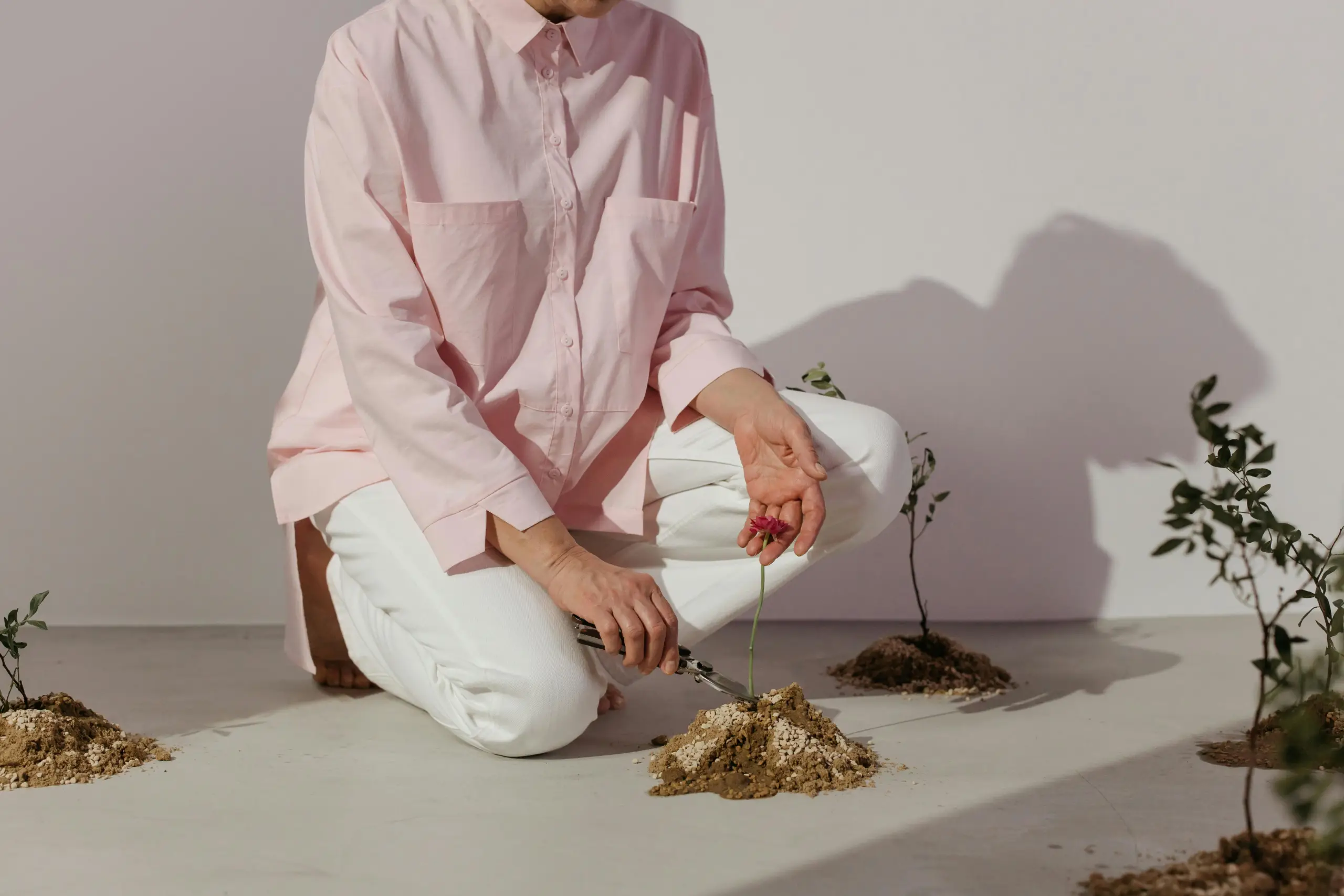 Person kneeling on a light floor, wearing a loose pink shirt and white pants, holding a small flower while tending to a cluster of soil mounds and tiny plants.