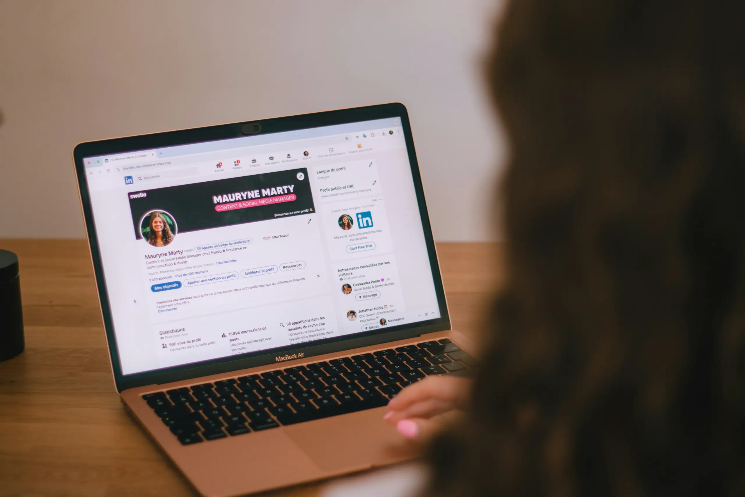 A person with long hair sits at a wooden desk, looking at a MacBook Air screen displaying a LinkedIn profile page of Mauryne Marty with a large banner and profile details.