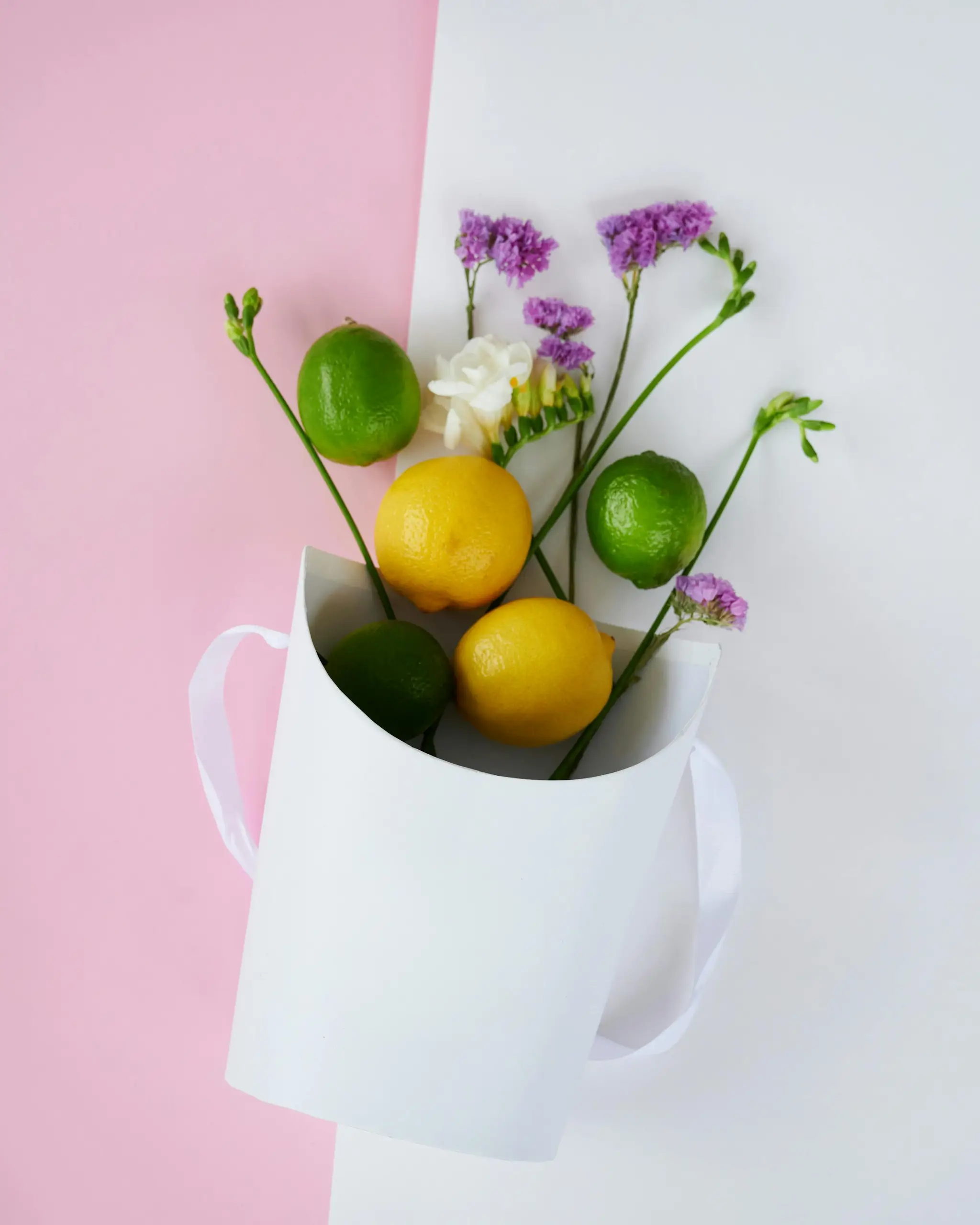 A white gift bag on a pink and white split background holds a bouquet of lemons and limes with purple flowers and a white bloom, arranged with green stems extending from the top.