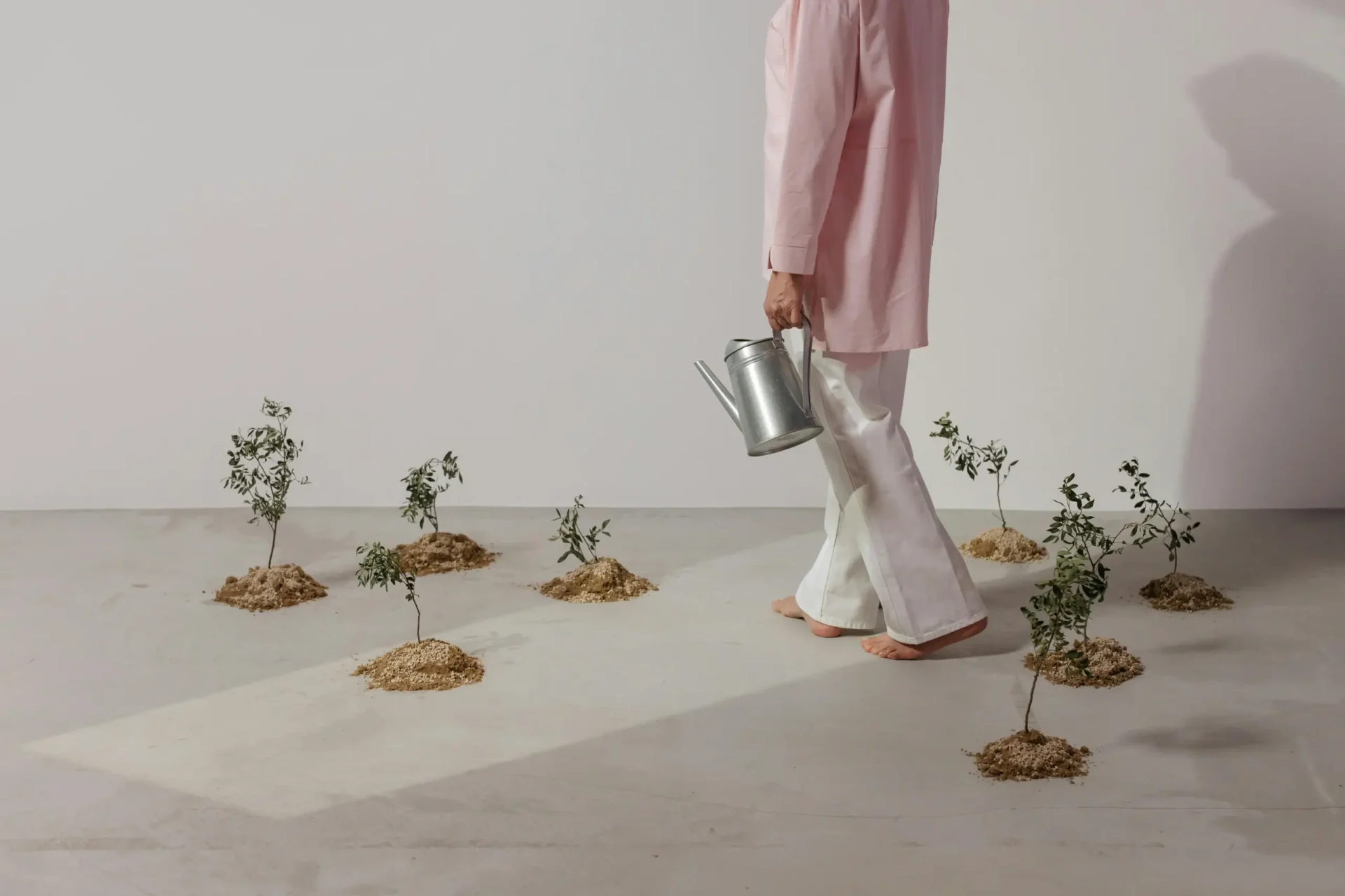 A person in a pink long-sleeve top and white pants walks barefoot across a light studio floor, carrying a metal watering can. Small plants in soil mounds are arranged around, creating a minimalist indoor garden scene.