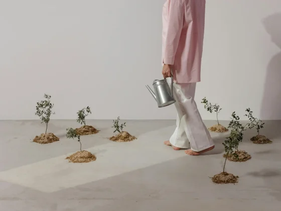 A person in a pink long-sleeve top and white pants walks barefoot across a light studio floor, carrying a metal watering can. Small plants in soil mounds are arranged around, creating a minimalist indoor garden scene.