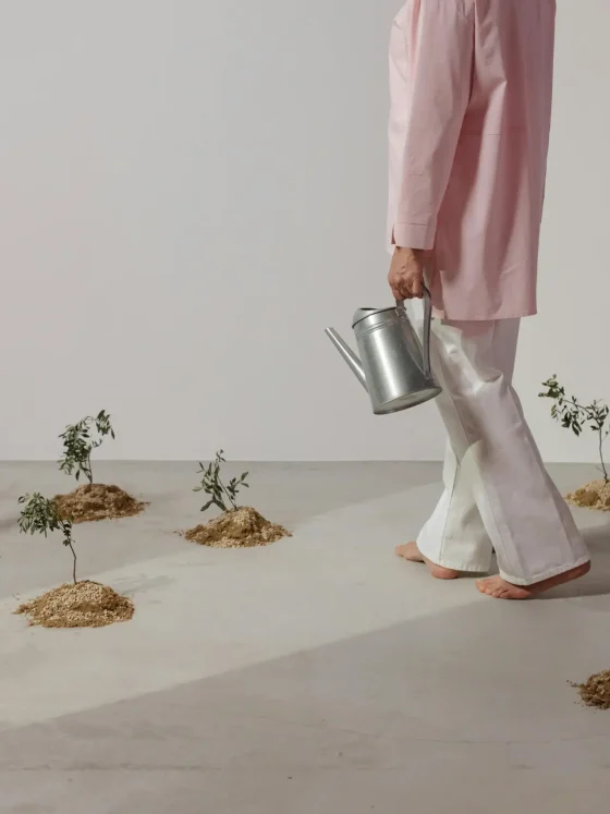 A person in a pink long-sleeve top and white pants walks barefoot across a light studio floor, carrying a metal watering can. Small plants in soil mounds are arranged around, creating a minimalist indoor garden scene.