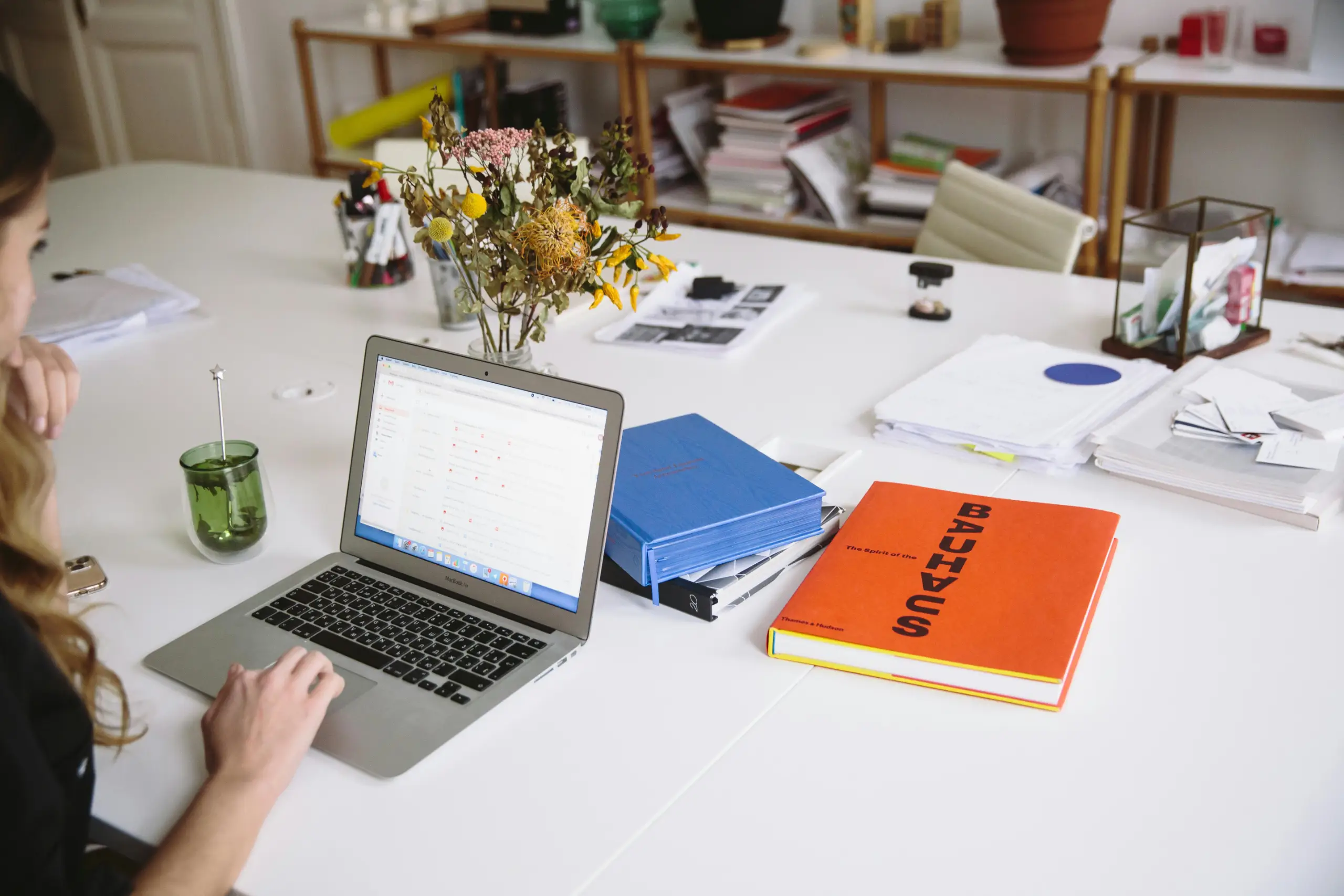 A white desk in a bright office scene with a silver laptop open to emails, a bouquet of dried flowers in a glass vase, and two colorful books including an orange one labeled 'BAU' beside a blue folder and stacks of papers.