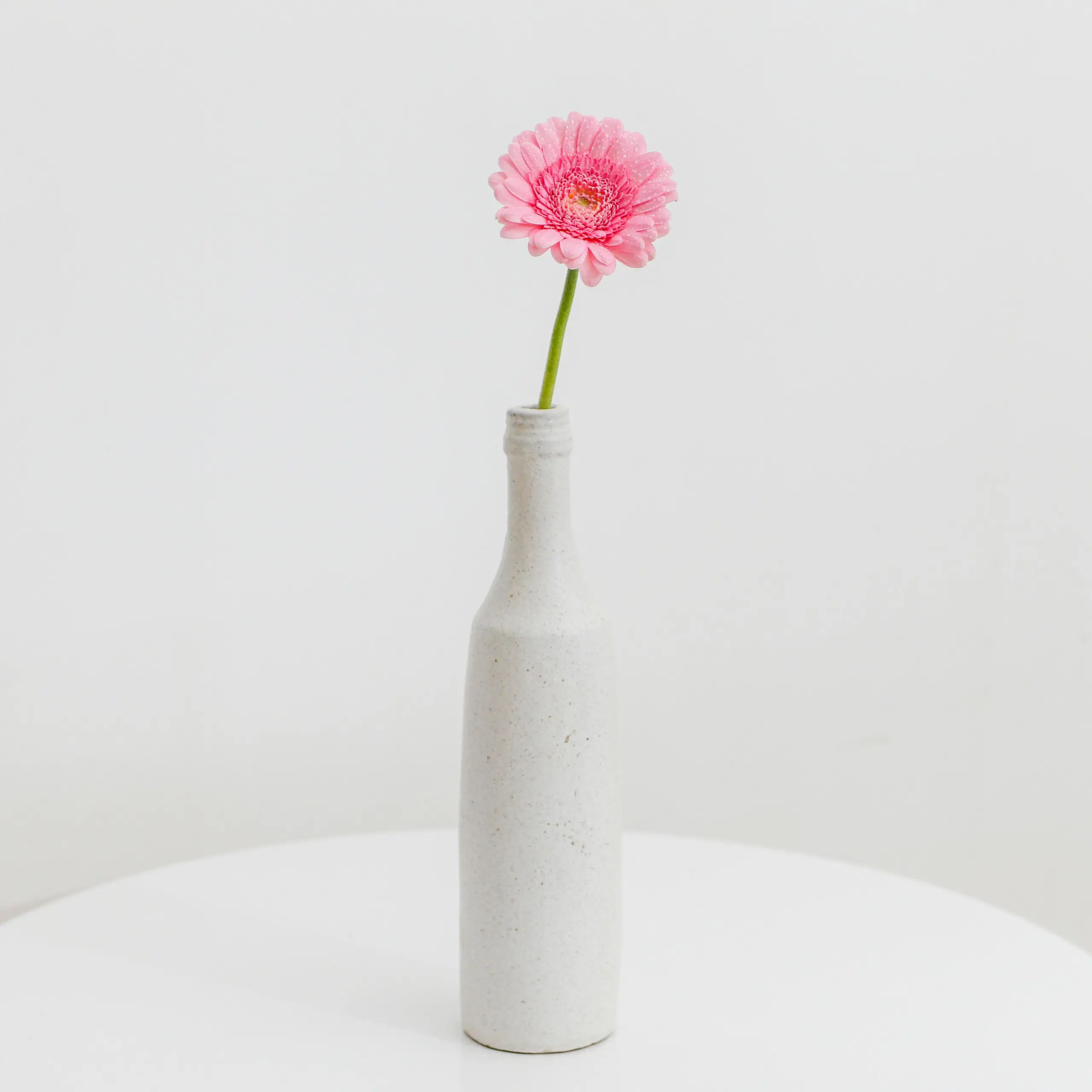A pink gerbera flower with a long green stem sits upright in a tall narrow white ceramic vase on a white circular table against a plain light background.