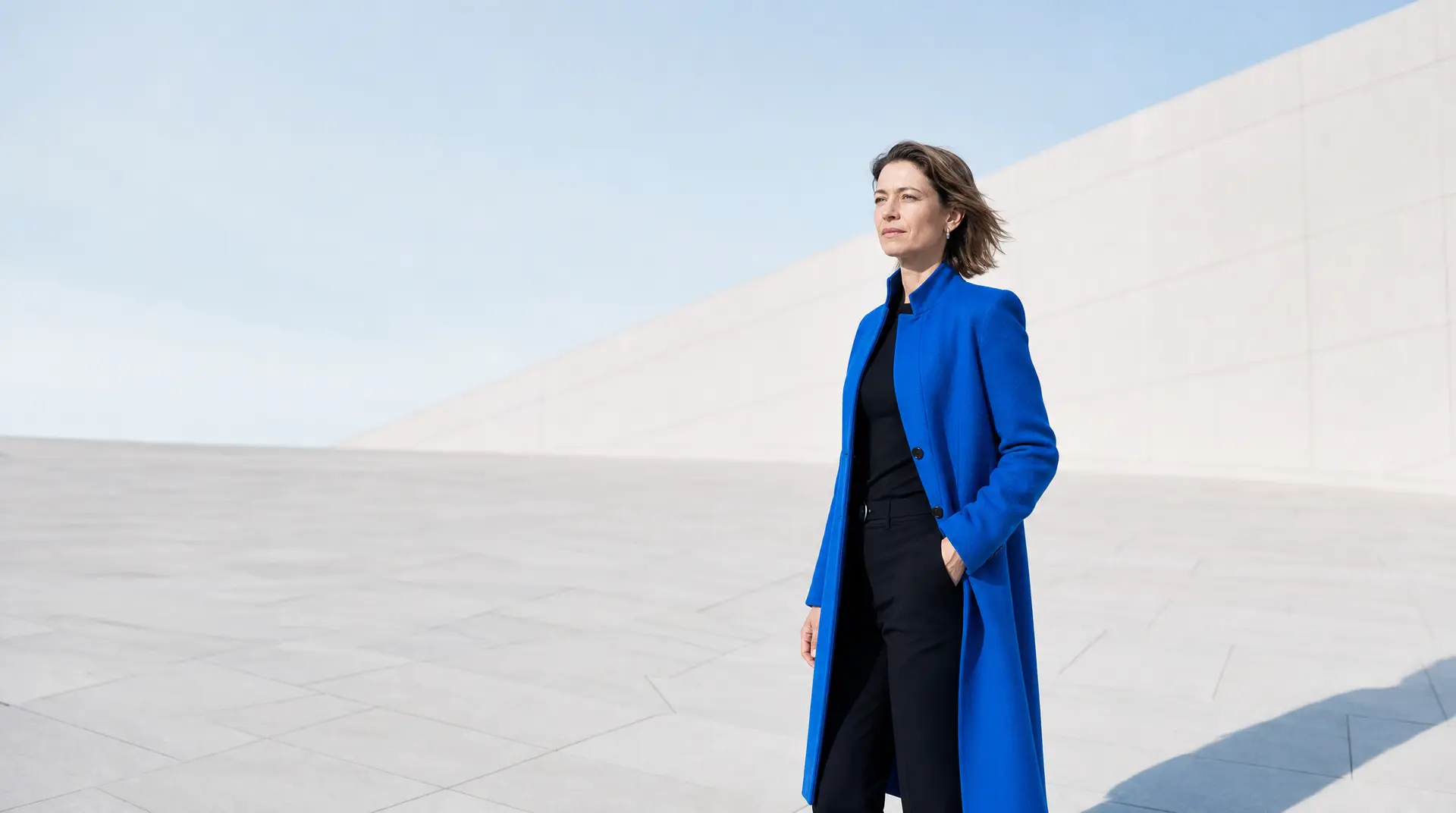 Businesswoman in a bright blue coat stands confidently beside a clean white stone wall, signaling personal brand linkedin.